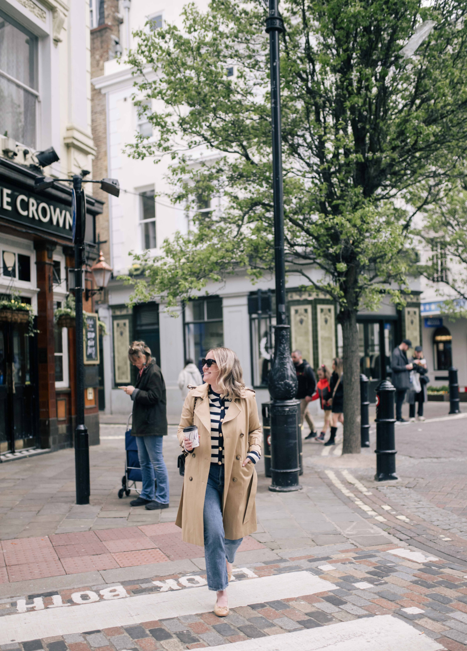 Meghan Donovan of wit & whimsy in a chic spring outfit featuring a striped cardigan, trench coat, and jeans, strolling through a London street.