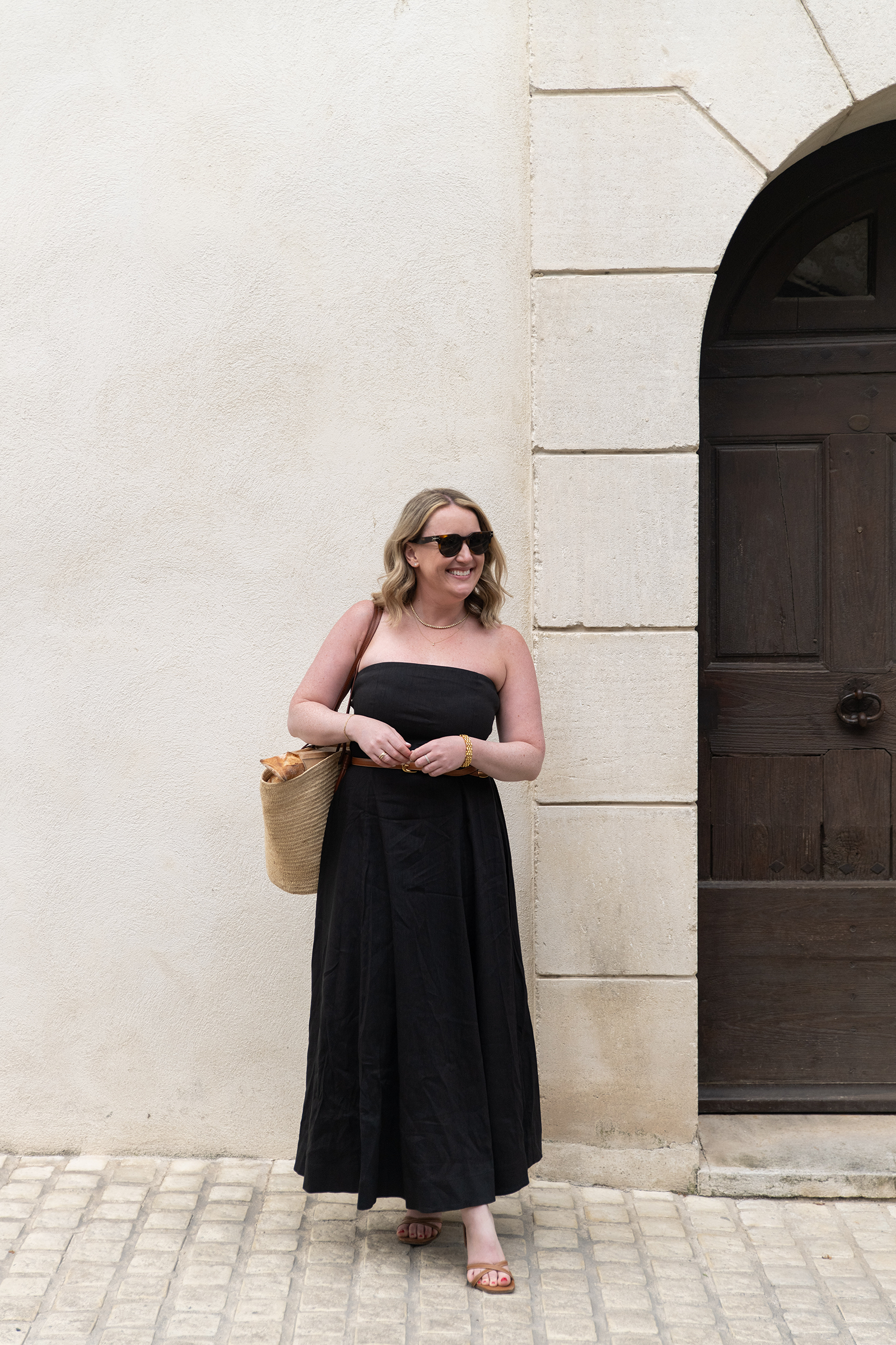 Woman in a strapless black maxi dress styled with brown leather slide sandals and a straw tote bag with baguettes, standing in front of a stone wall in Provence — an effortless example of what shoes to wear with black dresses in summer.