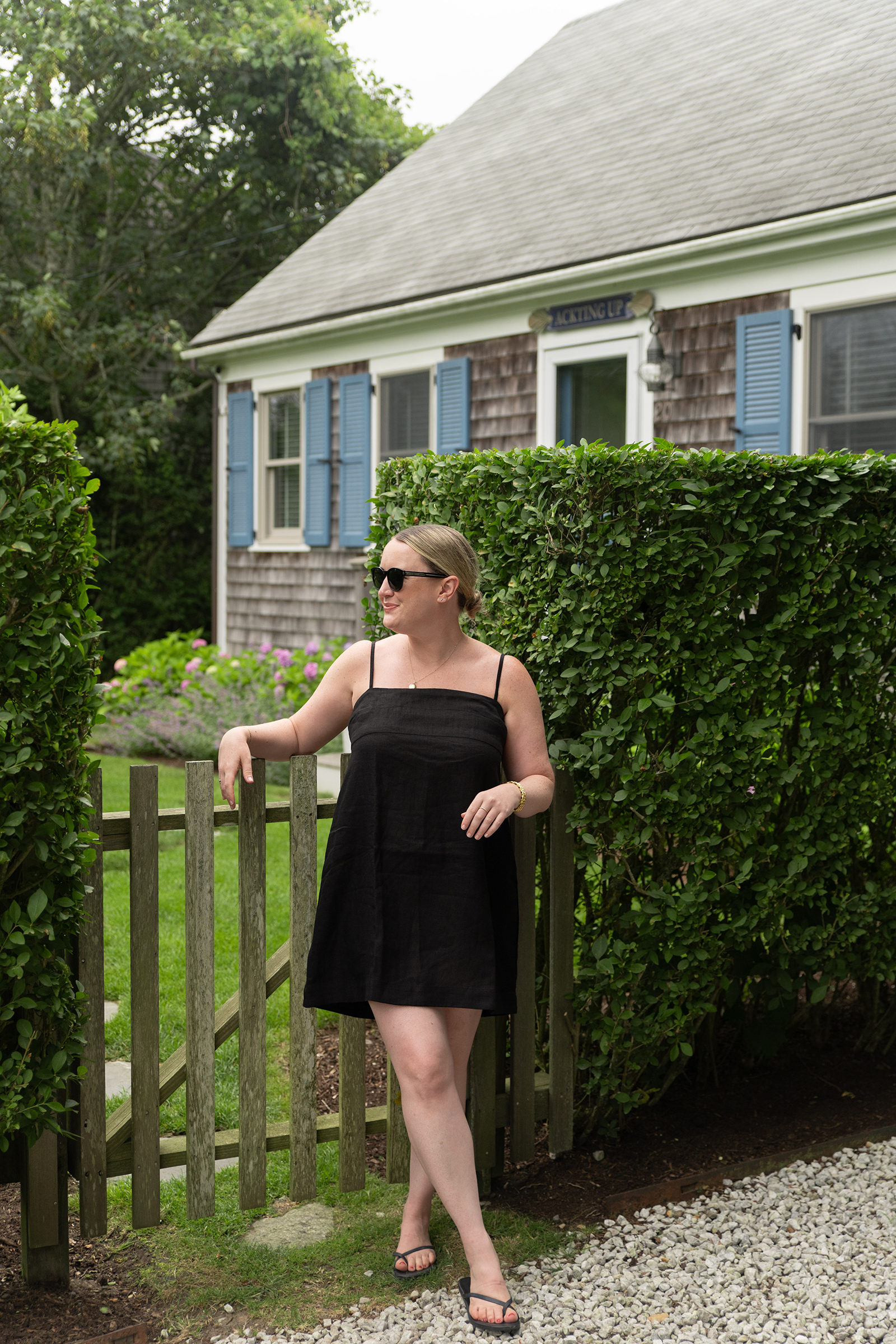 Woman in a black sundress styled with minimalist black flip flop sandals, standing by a garden gate in front of a Nantucket cottage — an easy summer take on what shoes to wear with black dresses. Woman in a black sundress styled with minimalist black flip flop sandals, standing by a garden gate in front of a Nantucket cottage — an easy summer take on what shoes to wear with black dresses.