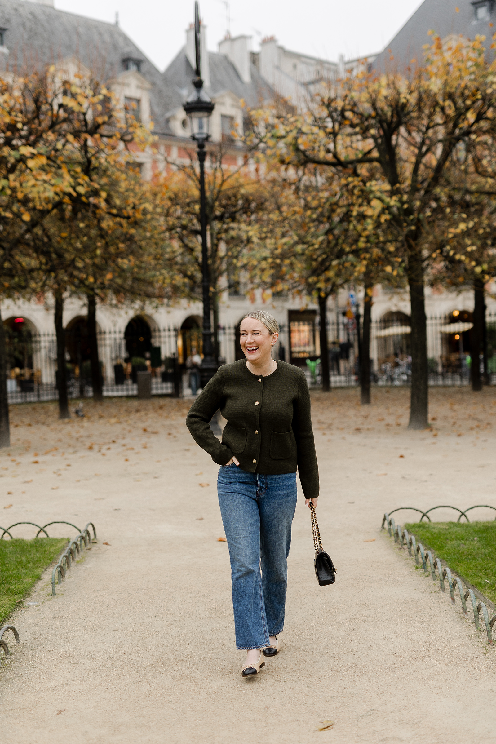 Woman in Paris wearing a dark green cardigan jacket, straight-leg jeans, and Chanel ballet flats, carrying a black handbag while walking through Place des Vosges in autumn.