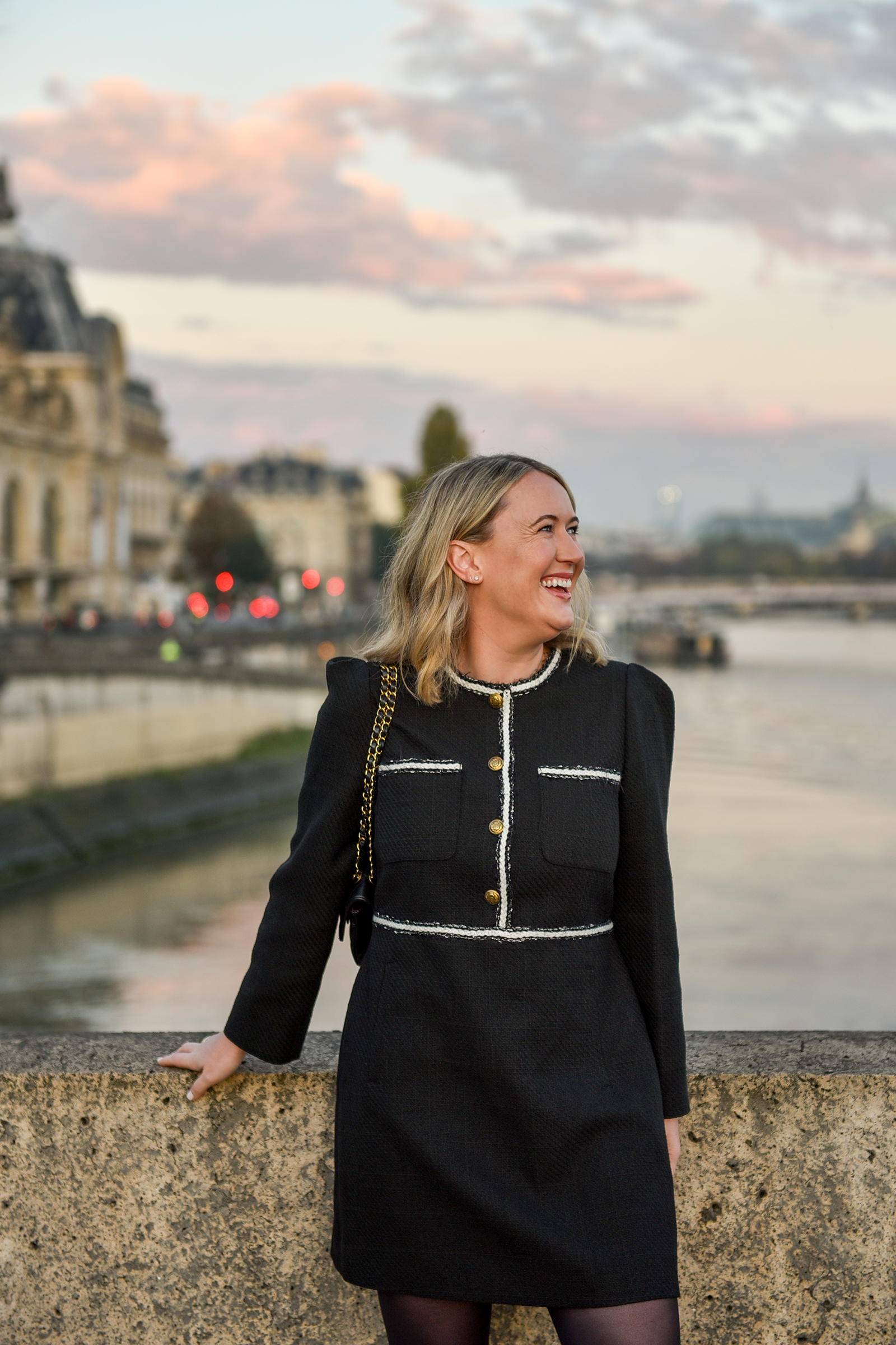 Woman in Paris wearing a black Me+Em tweed mini dress with gold buttons and white trim, standing along the Seine at sunset with city views in the background.