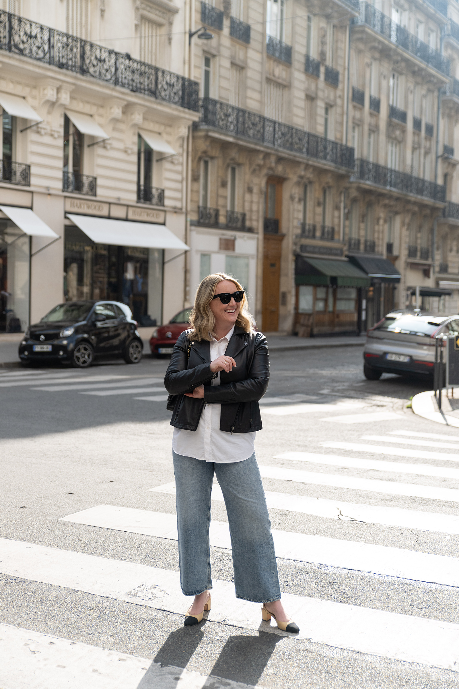 Woman wearing a black leather jacket, white button-down shirt, wide-leg jeans, and Chanel beige-and-black slingback heels, standing on a Paris street crosswalk.