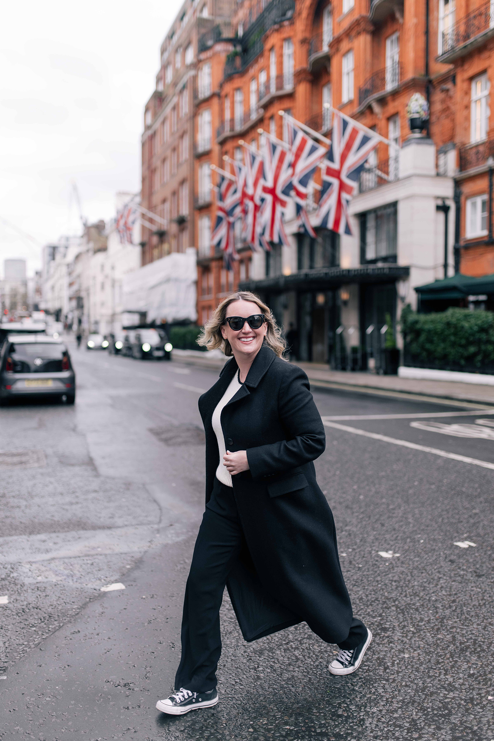Woman wearing a black coat, cream sweater, and black trousers with classic black Converse Chuck Taylor All Star low-top sneakers, walking across a London street with Union Jack flags in the background.