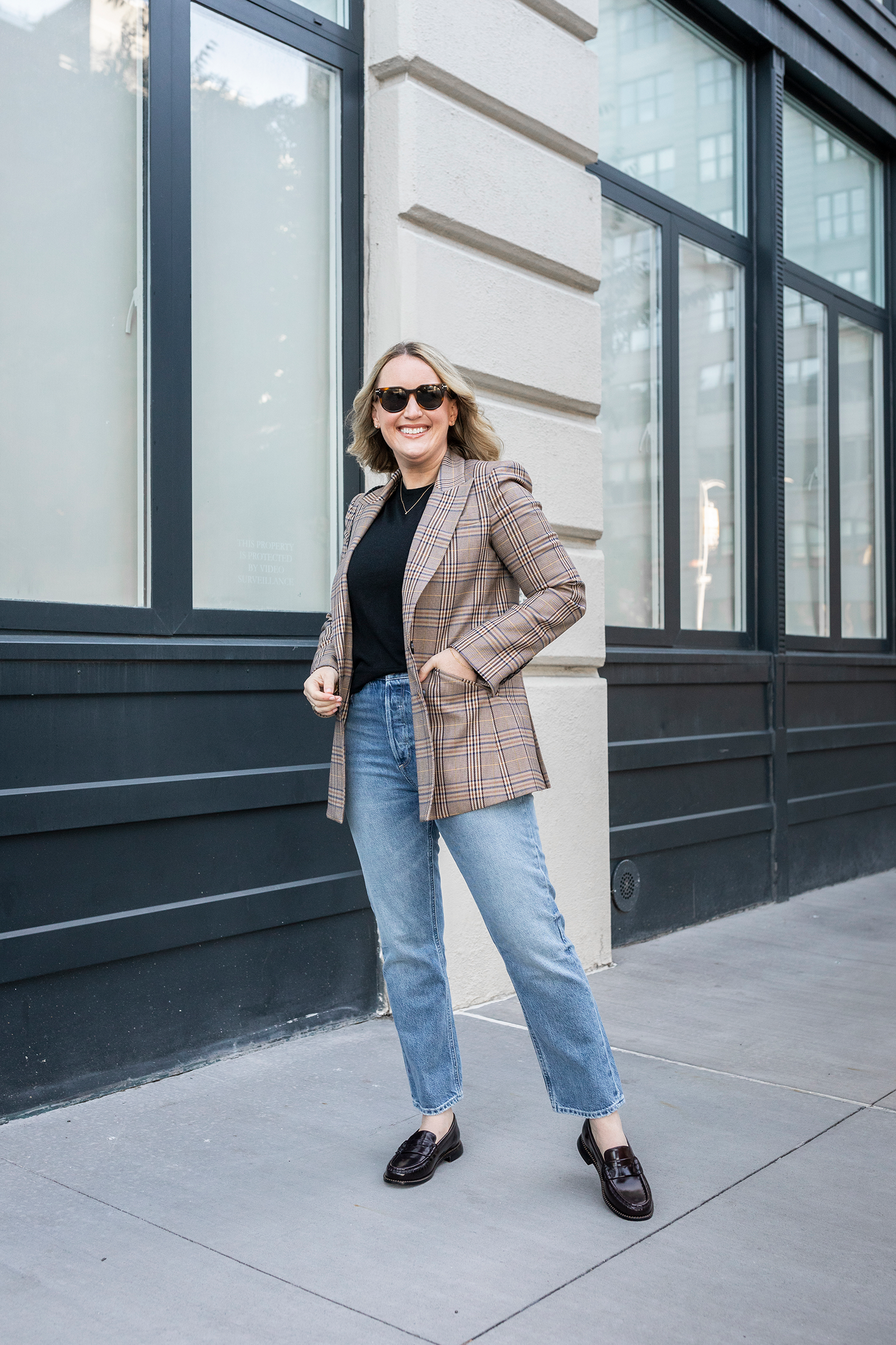 Woman wearing a brown plaid blazer over a black sweater with straight-leg jeans and classic black loafers, standing on a city sidewalk.