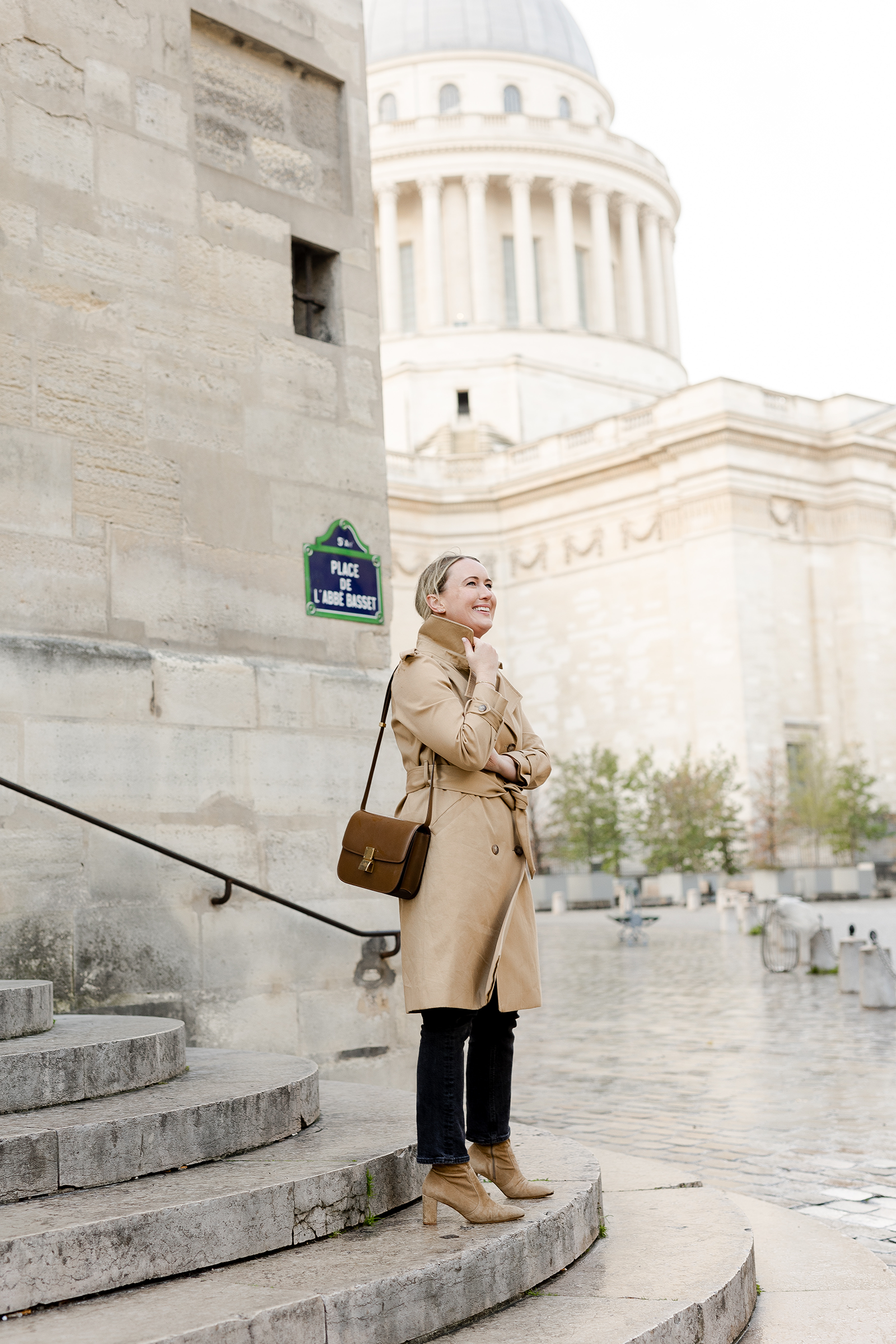 Woman in Paris wearing a classic Sezane beige trench coat tied at the waist, black skinny jeans, and tan block-heel suede ankle boots, standing on stone steps with the Panthéon in the background.