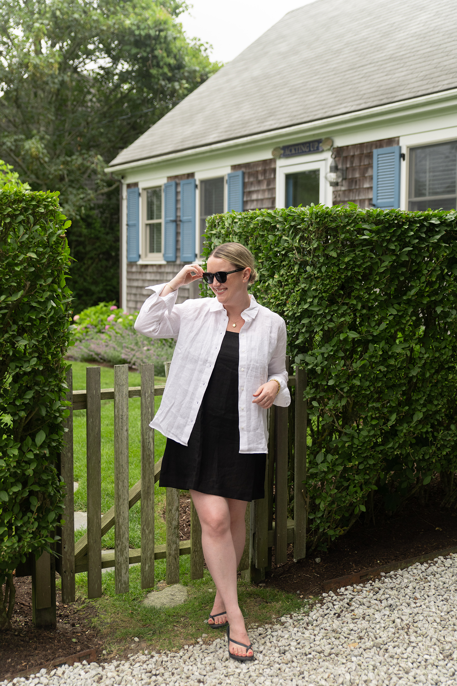 Woman in a black sundress layered with a white linen button-down shirt, wearing minimalist black flip flop sandals, standing by a garden gate in Nantucket.