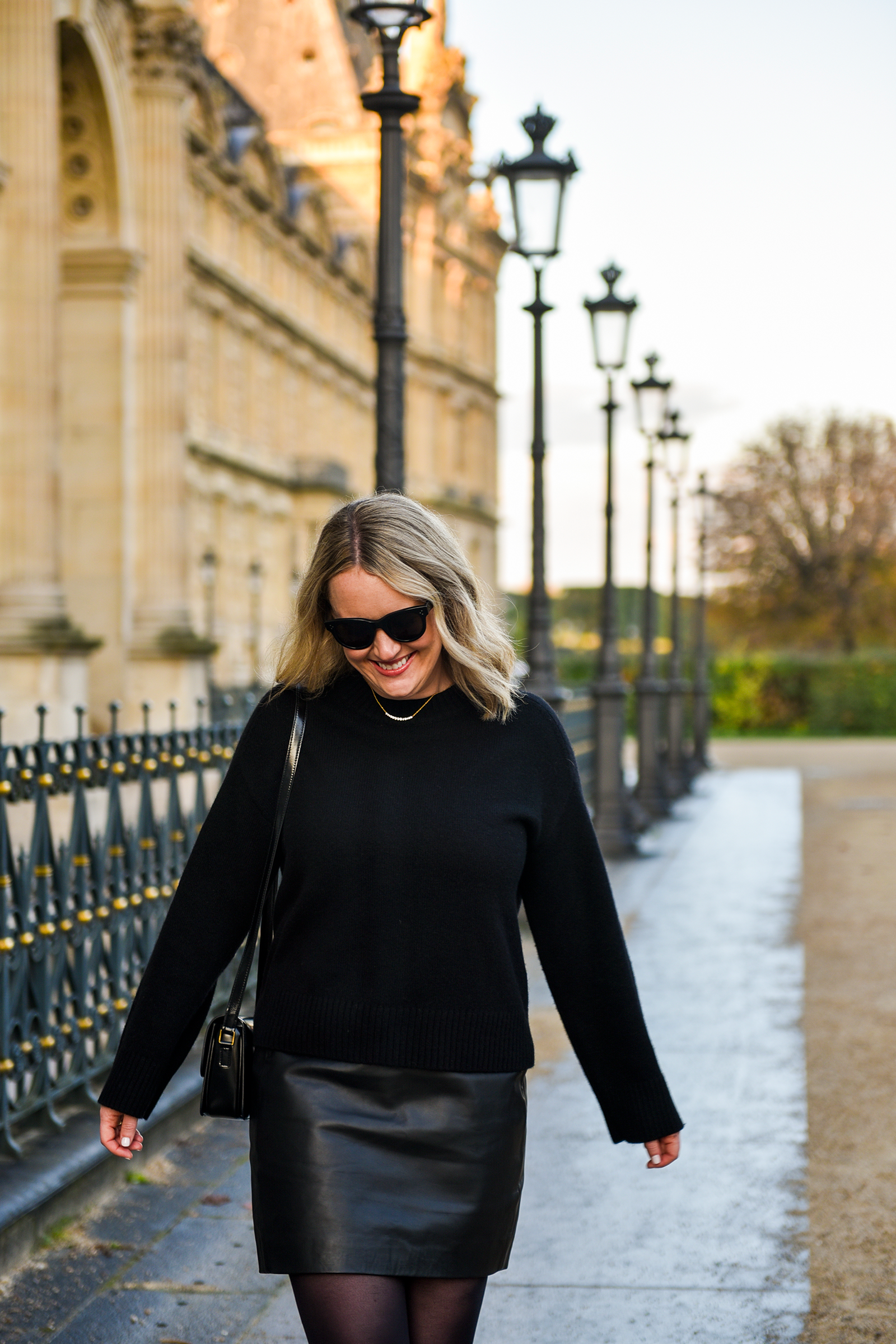Woman in Paris wearing a black sweater tucked into a black Sézane leather mini skirt with black tights and sunglasses, walking near the Louvre.
