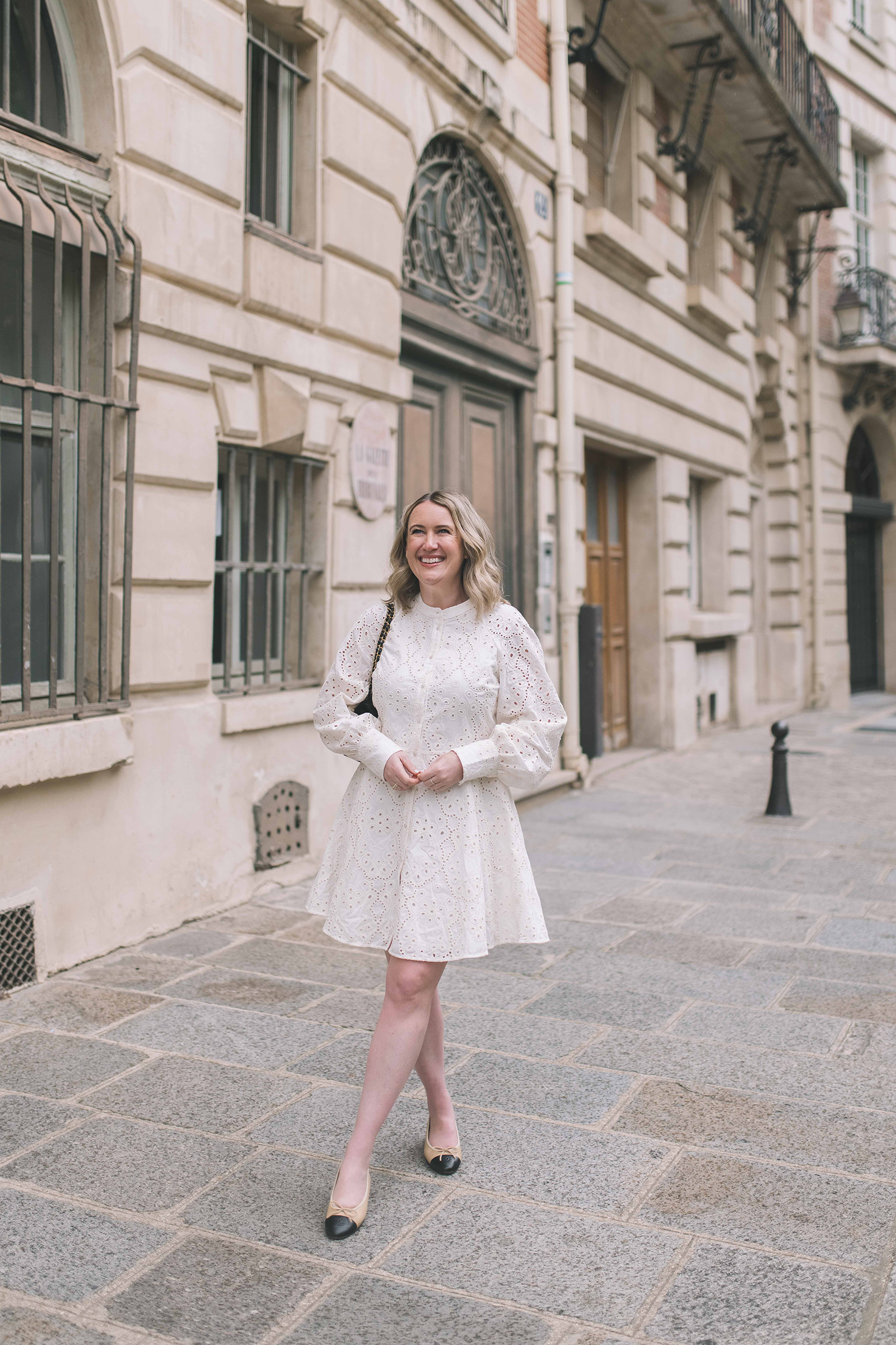Woman in a long-sleeve white eyelet mini dress styled with classic beige and black Chanel ballet flats, walking along a Paris street.