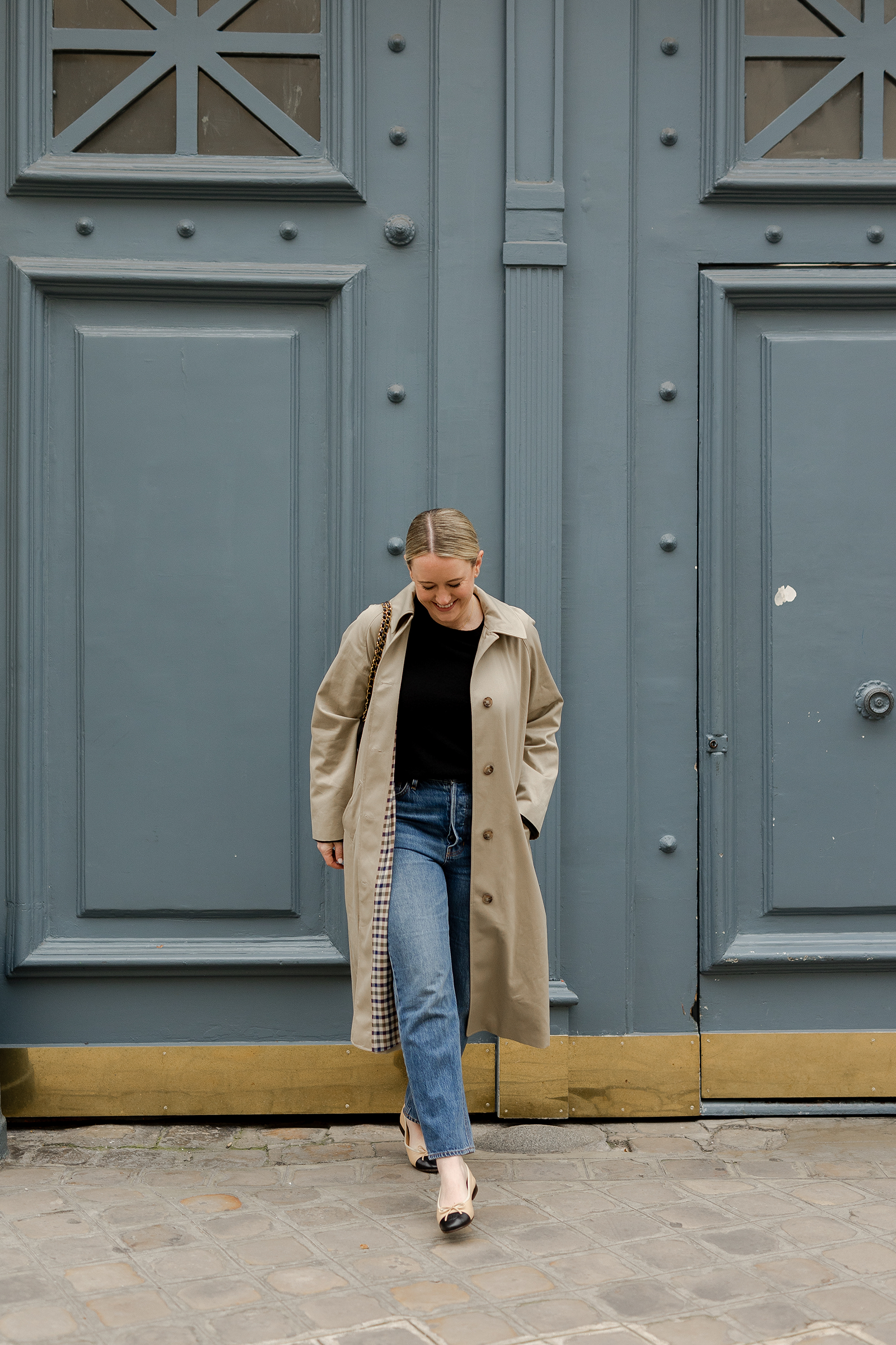 Capsule Wardrobe Basics Woman in Paris wearing a beige Sezane trench coat layered over a black top and straight-leg jeans, styled with Chanel two-tone ballet flats, standing against a blue French door.