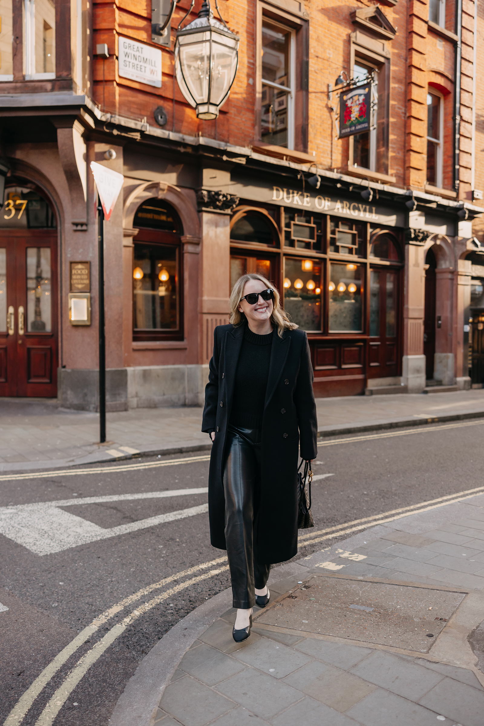 What to Wear in London Woman wearing black leather pants and a long black coat walking in London near a historic pub.