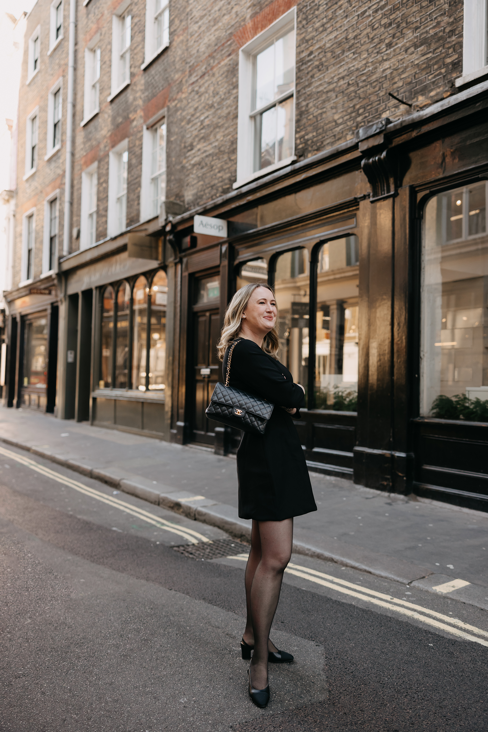 What to Wear in London Woman wearing a little black dress with sheer tights and black heels on a London street, styled with a quilted Chanel bag.