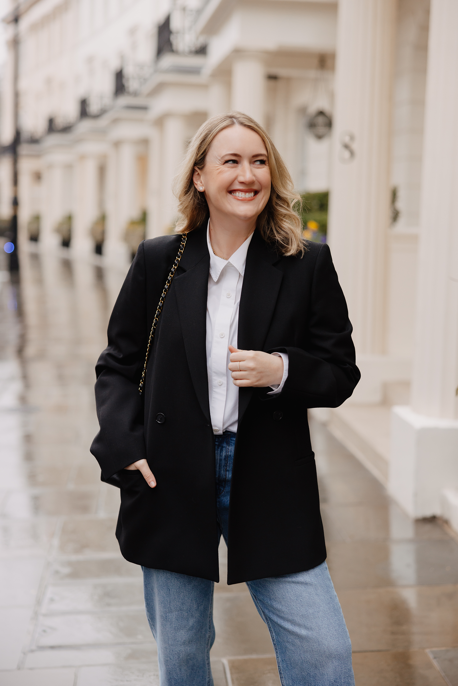 What to Wear in London Woman wearing a black blazer, white button-down shirt and blue jeans on a rainy London street.