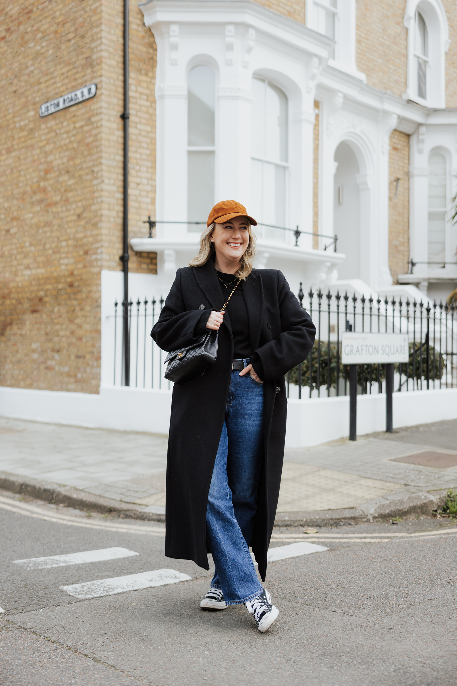 Fall Outfit Ideas With Jeans Woman wearing a black long coat, straight-leg jeans, sneakers, and a brown cap in London, showcasing a casual fall outfit for a London packing list