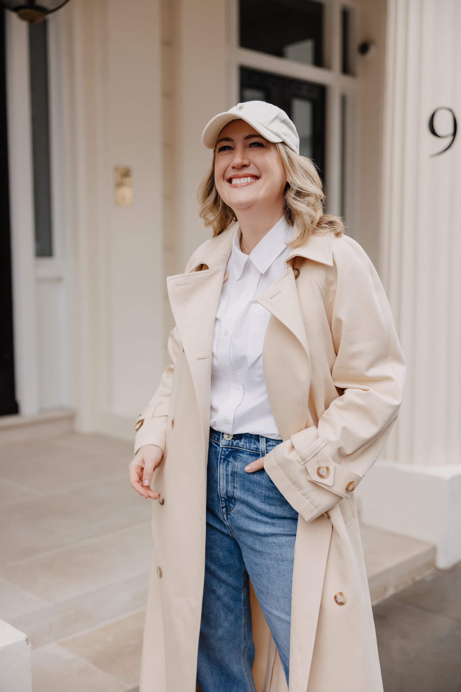 Woman wearing a beige trench coat, white button-down shirt, straight-leg jeans, and baseball cap in London, showcasing a chic spring outfit for a London packing list