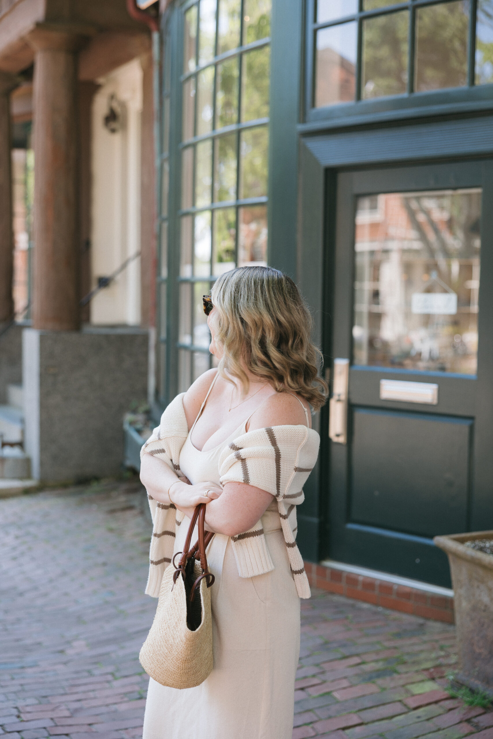 Woman wearing a cream knit tank dress with a striped cardigan draped over her shoulders and carrying a woven straw tote bag on a charming brick-lined street, demonstrating effortless spring sweater styling.