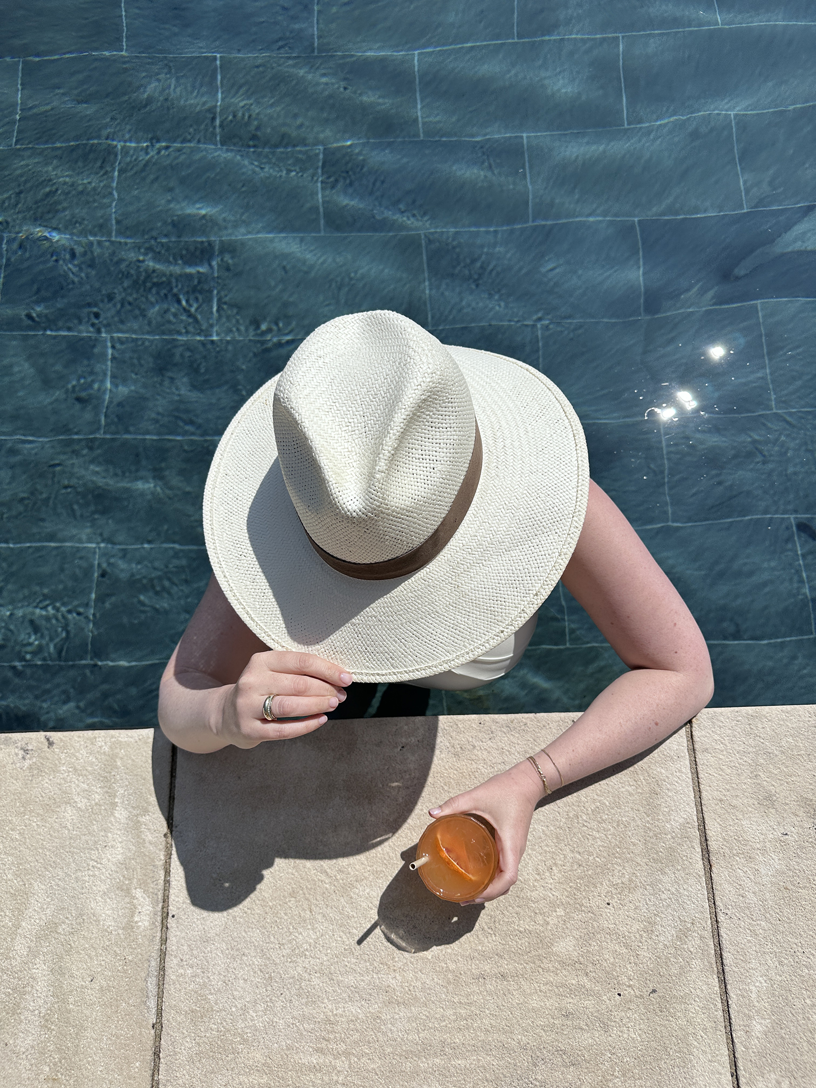 What to Pack for the Cotswolds Woman wearing a wide-brimmed straw hat and holding an Aperol spritz while relaxing by a pool on a sunny day.