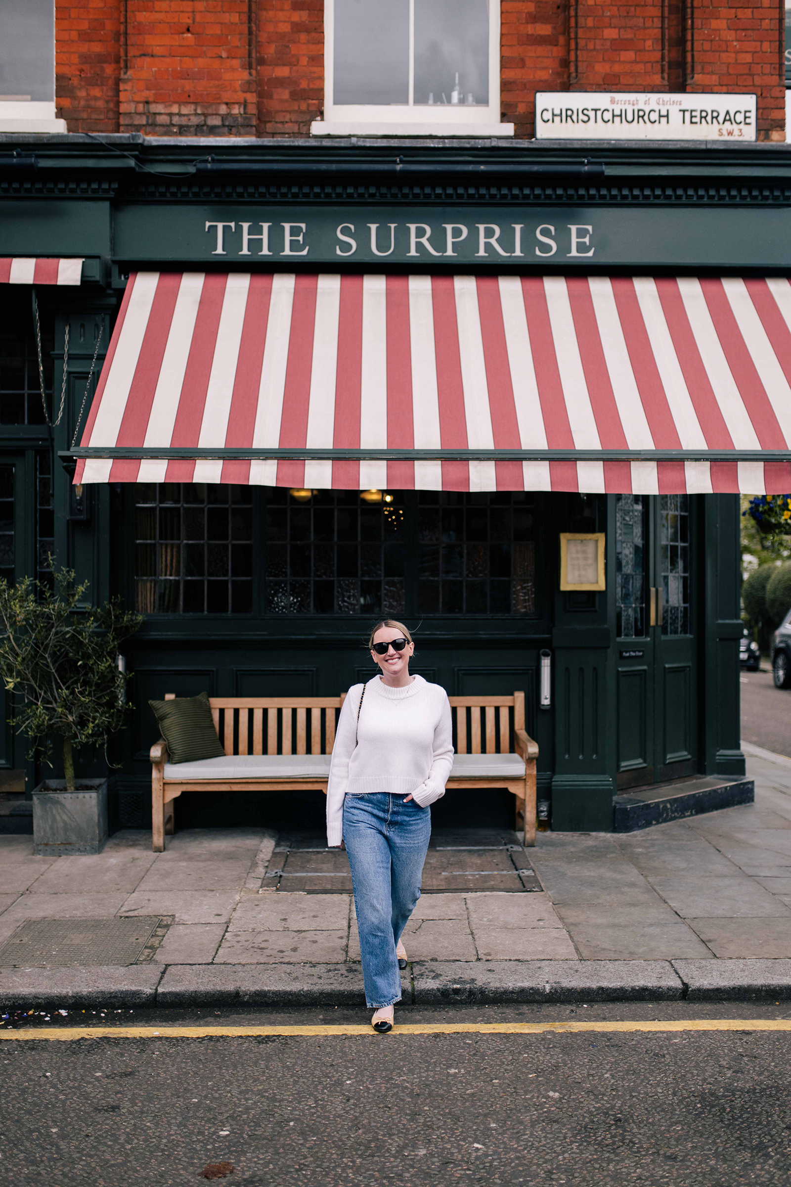 What to Pack for the Cotswolds Wit Whimsy wearing a white sweater, blue jeans, and cap-toe flats walking in front of The Surprise pub in London