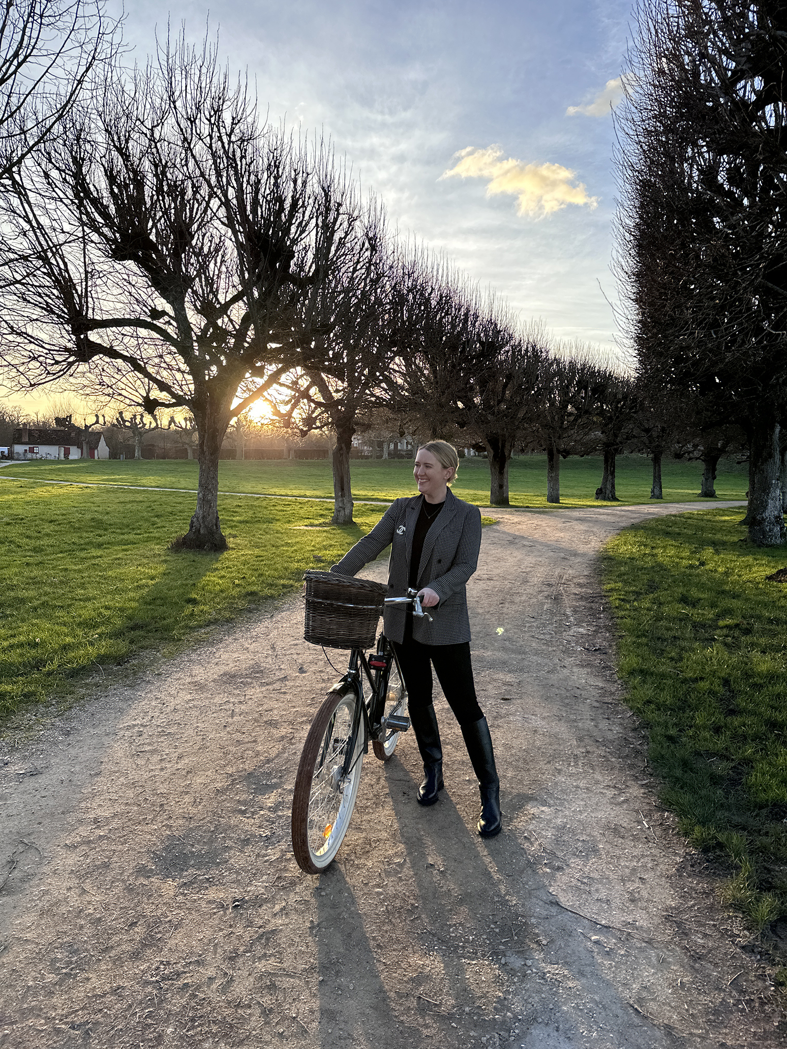What to Pack for the Cotswolds Woman wearing a plaid blazer, black turtleneck, and knee-high boots standing with a bicycle at sunset on a tree-lined path in the countryside