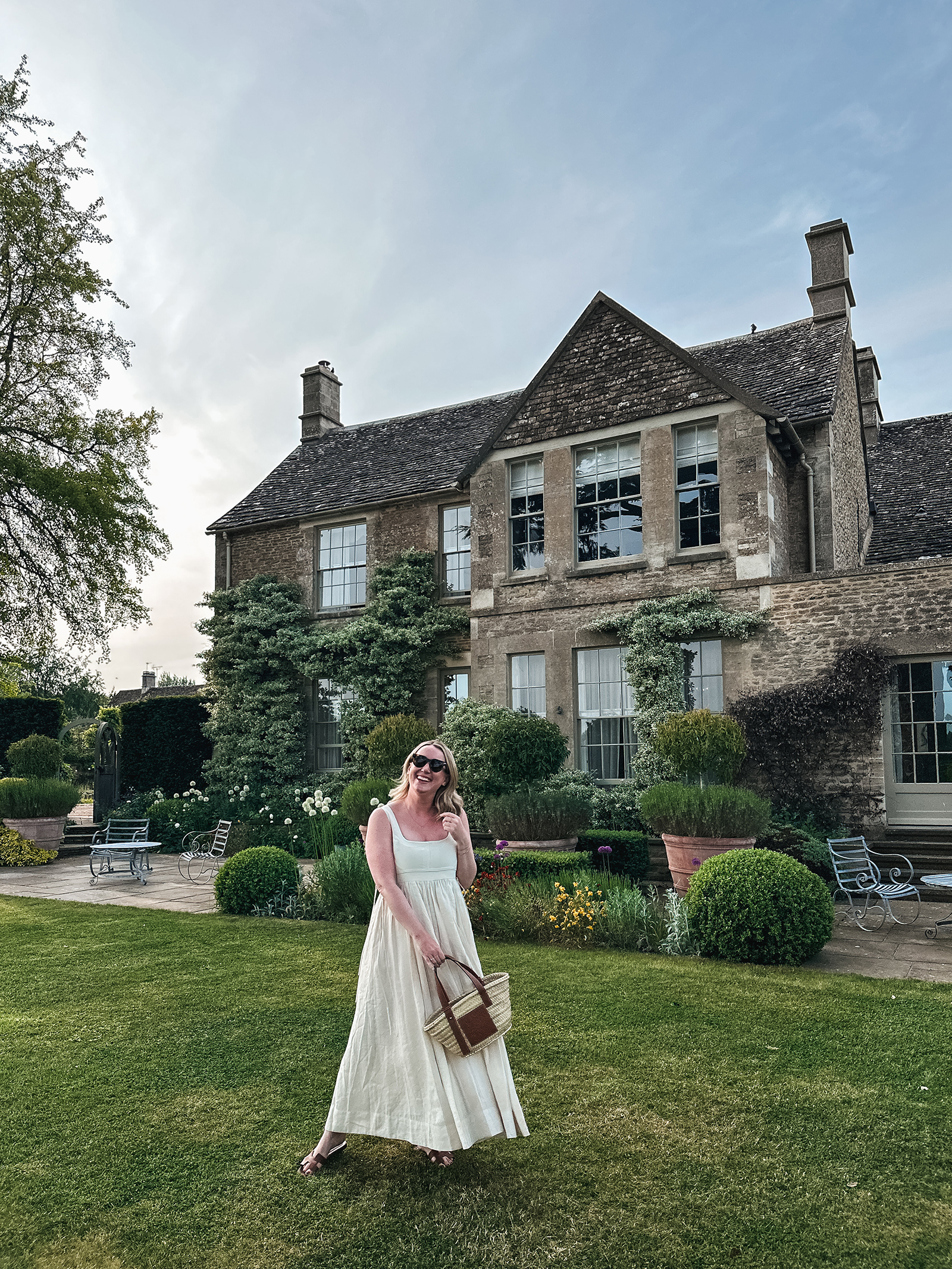 What to Pack for the Cotswolds Wit Whimsy wearing a white linen maxi dress and brown woven bag standing on the lawn in front of a charming stone house in the Cotswolds
