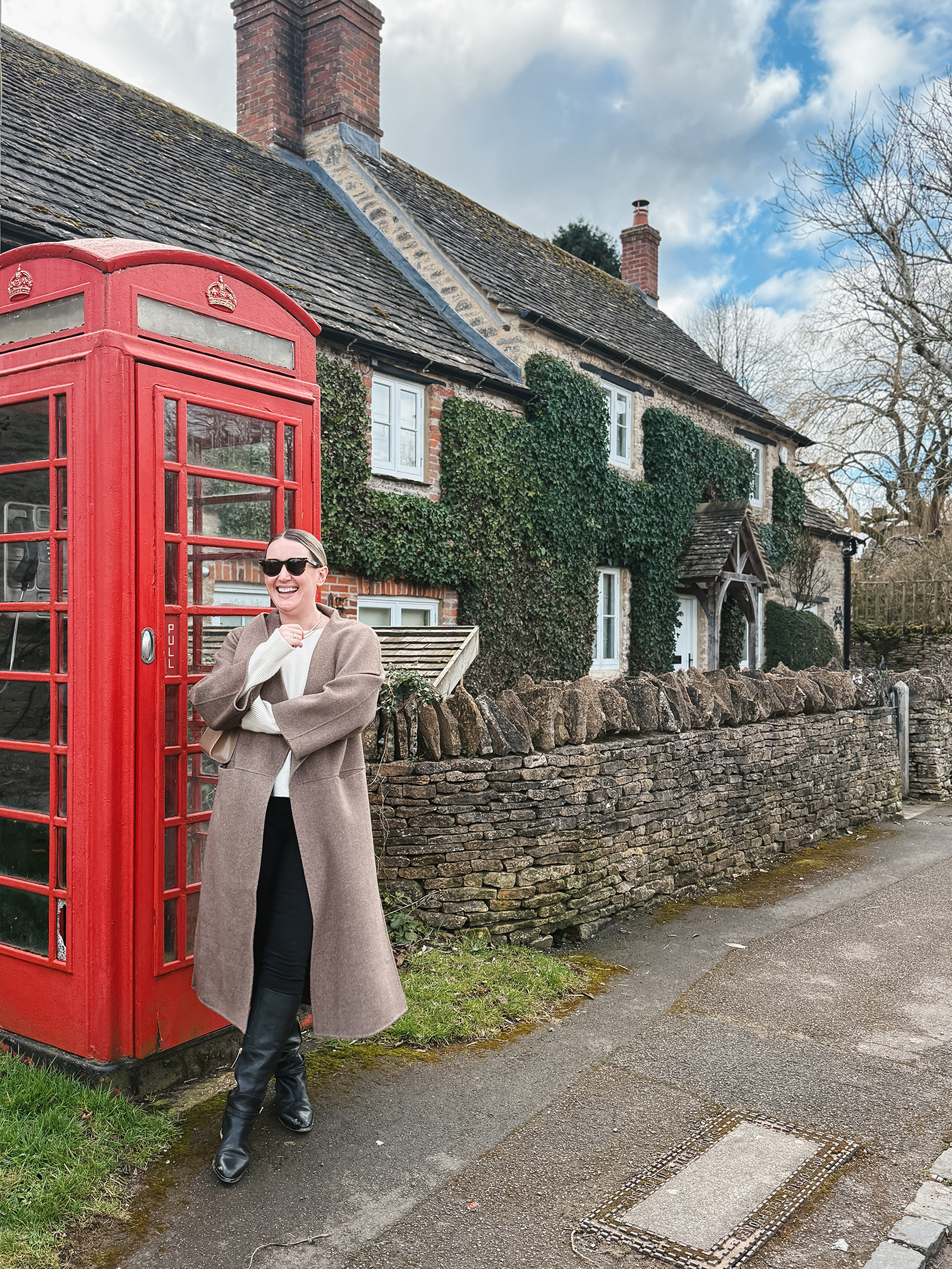 What to Pack for the Cotswolds Wit Whimsy wearing a long camel coat, white sweater, black jeans, and knee-high boots standing beside a red phone booth in front of an ivy-covered stone cottage in the Cotswolds